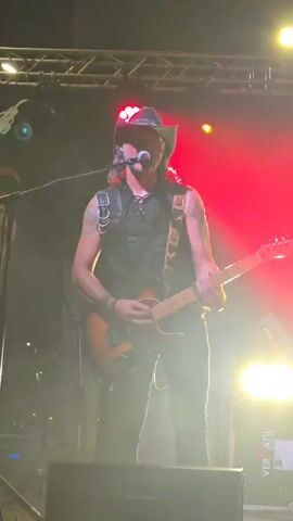 A man wearing a cowboy hat, black vest, and black pants plays a guitar on a stage. He is holding the guitar by its neck and strumming it with his right hand. His left hand is on the fretboard of the guitar. He is standing in front of a microphone and a speaker. There are lights on the stage, some of which are red. Another man, wearing a black shirt and ripped jeans, stands to the right of the guitarist, holding a bass guitar. There are speaker cabinets and stage equipment behind the musicians. The scene is dimly lit with stage lighting.