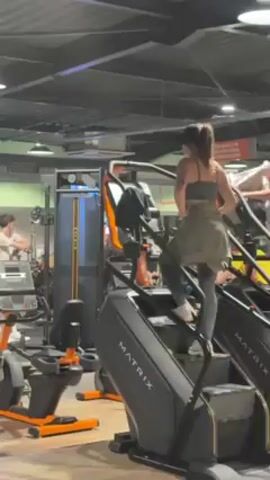 The video shows the interior of a gym. A woman wearing a sports bra, leggings, and a jacket tied around her waist is using a Matrix stair climbing machine. Other gym equipment is visible in the background, including treadmills, an elliptical machine, and a weight machine. Another person can be seen on a treadmill in the background as well.  The gym has a black ceiling with fluorescent lighting.