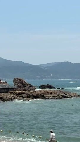 A person wearing a white hat and a white top is walking on a beach towards the ocean. The person is walking away from the camera. The ocean has choppy waves and is a greenish-blue color. There are several large rocks in the ocean near the shore. In the background, there are green, hilly mountains under a clear, blue sky.