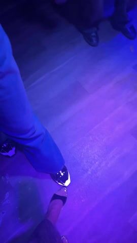Two people are standing on a wooden floor that is lit with shifting colorful lights. One person is wearing gray pants and black sneakers with white accents. The other person is wearing a patterned garment, and black heeled sandals. The lighting changes from red to purple to yellow and green to blue, highlighting the movements and shapes in the scene. The shadows of the people are visible on the floor.