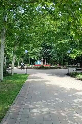 This video pans across a park on a sunny day. The scene features lush green trees, a paved walkway, park benches, and decorative landscaping. In the background, there's a circular monument with a statue and a large sign displaying the numbers "100," adorned with various images. One person is seen cycling on the path, and another person is sitting on a bench. The park appears to be a public space, possibly a town square or a recreational area, with an outdoor stage visible in the distance. The overall atmosphere is peaceful and leisurely, suggesting a pleasant day outdoors. No specific time of day is indicated, but the bright sunlight suggests daytime. The location is identified as Severskaya, Russia.