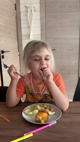 This is a vertical, eye-level, medium shot video of a young girl sitting at a wooden table, eating sliced apples from a white plate. The girl has blonde hair and is wearing an orange t-shirt with a cat graphic and a pink beaded necklace. She holds a fork in her left hand and brings apple slices to her mouth with her right hand, sometimes directly from the plate. A half-eaten apple is prominently placed in the center of the plate. Two markers, one pink and one yellow, lie on the table in front of the plate. In the background, a white macrame wall hanging is visible on a light-colored wooden wall. Two wooden doors with black handles are also in the background. The lighting suggests it is daytime, likely indoors in a home setting. The girl's expression is playful and engaging as she eats.