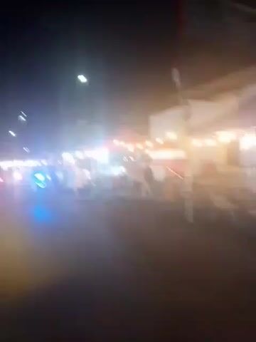 The video shows a street at night with several food stalls set up. A motorcycle with bright blue lights is in the foreground, moving from left to right. There are people at the food stalls, some of whom appear to be customers or vendors. The stalls are illuminated by string lights. A parking sign is visible on a pole in the background. The overall scene suggests a bustling night market or street food area.