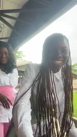 Two young women are positioned in the foreground. The woman on the right has long, braided brown hair and is wearing a white collared shirt with a light green corset over it. She is smiling and making a peace sign with her left hand, while her right hand is near her face holding a pink object, possibly a phone. The woman on the left has long, dark, wavy hair and is wearing a white collared shirt with a pink sash around her waist. She is looking towards the right and smiling. They appear to be interacting closely, with the woman on the left touching the right woman's hair or clothing. The background suggests an outdoor or semi-outdoor setting with a covered roof structure above them and foliage visible in the distance. The lighting is bright, indicating daylight. Other people are partially visible in the background.