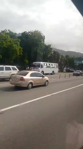 A static shot taken from a vehicle moving on a highway shows a gold car driving in the adjacent lane. The car is seen from its rear, and as the vehicle with the camera moves, the car gradually disappears behind a concrete barrier. The background features trees, buildings, and mountains under a cloudy sky. Other vehicles are visible on the highway. At the edge of the frame, part of someone's head can be seen.