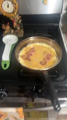 A video shows a black gas stove. On one burner is a stainless steel frying pan with eggs and bacon cooking in it. On another burner is a stainless steel pot of cooked food with a spoon in it. There is a ceramic spoon rest on the stovetop.