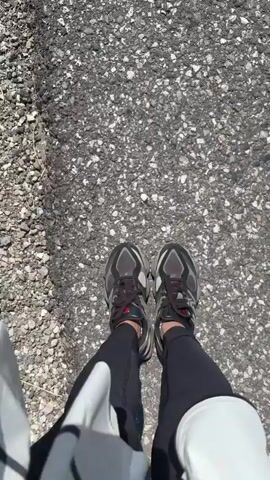 The video begins with a close-up of a person's feet in athletic shoes walking on a paved surface. The camera then pans up to show the person walking on a path, and then shifts to a wider view of the path. A person and a dog are walking on the path, and in the background, two other people are walking ahead. The path is surrounded by green grass and rolling hills in the distance, with mountains visible on the horizon under a clear sky. The camera continues to follow the people and the dog as they walk.