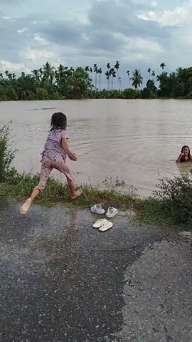 A video shows a group of children playing in a body of water next to a road. The video begins with a child in a patterned dress jumping into the water. Another child in a gray t-shirt then splashes into the water, followed by a child in a green outfit. Two pairs of sandals are visible on the road next to the water, and a motorcycle is parked on the right. Palm trees and other vegetation line the far bank of the water. The sky is cloudy.