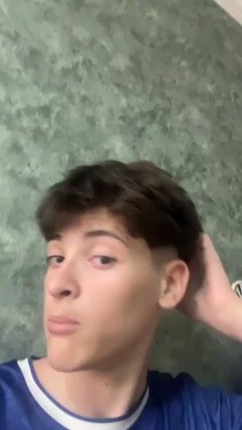 The video shows a young man with dark, styled hair and a blue shirt. He is touching his hair with his right hand and looking towards the camera. The background is a textured green wall.