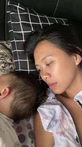 The video captures a close-up, high-angle shot of a woman and a child resting together. The woman, with long dark hair, is lying on her side, initially with her eyes closed, suggesting sleep or deep rest. She is wearing a light-colored top, possibly with a faint pattern. Beside her, a young child with light brown hair is also lying down, seemingly asleep, with only the back of their head and ear visible. The child is partially covered by a white sheet or blanket with small colored dots.

The setting appears to be a bedroom, indicated by a black headboard and a black and white checkered pillow behind the woman. Another patterned sheet or blanket is visible beneath the child. The lighting suggests it is daytime. At the 0:02 mark, the woman briefly opens her eyes and looks forward before closing them again. Both subjects appear peaceful and calm. There are no visible texts or specific location cues within the frame to confirm the provided context of Ngasinan, Indonesia.