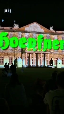 The video captures an exterior night scene of a building illuminated with a projection. A large, glowing green text that reads "Goedfner" is prominently displayed across the facade of the building. The building appears to be a classical structure with columns and windows. The lower portion of the building's facade is illuminated with yellow light, creating a contrast with the green text. In the foreground, a group of people are seated on the grass, facing the building, appearing to watch the projection. The overall environment suggests an outdoor event or performance.