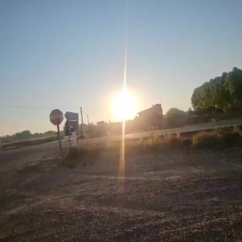 The video shows a sunrise over a rural landscape. The sun is a bright, hazy orb in the sky, casting a golden light over the scene. In the foreground, there is a gravel road with a stop sign and a traffic signal. In the background, there are buildings and trees. The sky is clear and blue, with no clouds visible. The overall impression is one of peace and tranquility.