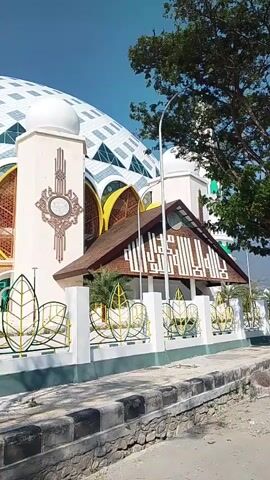 The video displays a wide shot of a mosque's exterior on a clear day. The mosque features a large white dome adorned with a geometric pattern and intricate window designs. A minaret stands to the right of the dome. In front of the mosque, there is a white fence with decorative leaf motifs. The surrounding environment includes a sidewalk and some greenery. The camera appears to be stationary.