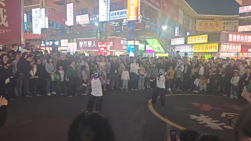 In a nighttime outdoor scene, two performers with illuminated face coverings are dancing in a marked circular area. They are wearing matching white t-shirts with graphics and baggy black pants. The circular area is located in a street or plaza surrounded by a large crowd of onlookers of various ages. The crowd includes adults and children, most of whom are standing and watching the performance. Some individuals are holding up phones, presumably to record or photograph the event. Behind the crowd are buildings adorned with numerous bright, colorful signs written in Chinese characters. The signs indicate the presence of various businesses and shops. The overall atmosphere appears to be lively and festive, with a public performance attracting a large audience in a commercial area.