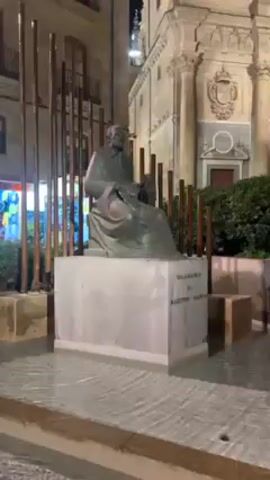 The video shows a statue of a man sitting on a pedestal, surrounded by a shallow pool of water. The water is gently flowing, creating a ripple effect. The statue is made of a dark metal, and the pedestal is made of a light-colored stone. The surrounding area appears to be a plaza or courtyard, with buildings visible in the background.  The video pans across the surface of the water, showcasing its movement and the reflection of light. The overall scene is calm and serene.