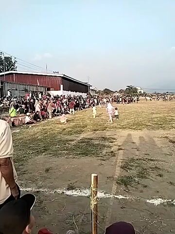 A wide outdoor shot shows a crowd of spectators sitting on the ground along the edge of a grassy field. In the background, a long building with a red roof and white walls is visible. Several people are standing and walking in the field, including children. Two children are in the foreground, facing away from the camera, watching the activity in the field. One child is wearing a tan shirt and pants, and the other is wearing a red shirt with a Spider-Man design. The sky is clear and bright, suggesting daytime. The ground in the foreground appears to be dirt with some dry grass.
