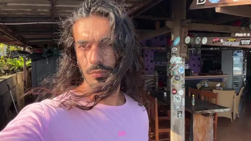 The video is a selfie taken by a man with long, dark, wavy hair and a beard, wearing a pink t-shirt. He is standing outdoors in what appears to be a beach bar or restaurant. The background shows a wooden structure with tables and chairs, a bar area with shelves stocked with bottles, and various decorations including stickers on a pillar. The lighting suggests it is daytime, possibly late afternoon, with sunlight casting shadows. The man is looking directly at the camera. The video is shot from a selfie perspective, with the man holding a phone or camera. He turns his head slightly to the right and then back to the camera. The environment appears casual and relaxed.