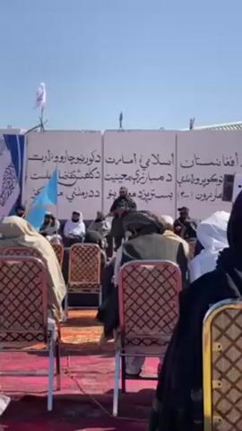 The video captures an outdoor event where a person is speaking at a podium in front of a seated audience. The speaker, dressed in what appears to be military attire, stands behind a banner written in Arabic. The audience, comprised mainly of men wearing traditional clothing, sits in chairs arranged on a red rug. A small flag with a turquoise emblem is visible on the side. The background includes what appear to be tents, with a clear blue sky overhead.