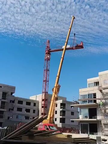 A construction site with two cranes and unfinished buildings under a blue sky with scattered clouds. One crane is red and tall, while the other is yellow and mobile. The yellow crane is lifting a section of construction material. A red van is parked near the base of the yellow crane. Several workers are present, one of whom is visible on scaffolding. Piles of lumber are in the foreground.