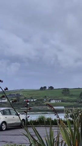 The video shows a landscape view with a cloudy sky. In the foreground, there is a silver car parked on the left side. Further back, there is a green hill with houses and trees. The ocean is visible in the middle ground. There are also some plants in the foreground, partially obscuring the view. The video pans to the right, revealing more of the landscape and a dark colored convertible car.