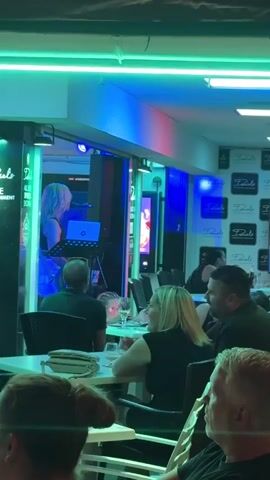 A woman with blonde hair stands at a podium with a laptop and microphone, performing. The scene is an outdoor seating area of a bar or restaurant illuminated by neon lights. Several tables with people are visible, some seated and others standing. The bar has "COCKTAILS" written in large letters above it.