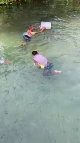 Three young children are in a shallow river. One child is holding an empty white bucket above the water. Another child is swimming. The third child is standing waist-deep in the water. The river water is clear enough to see the rocky bottom. The river banks have green foliage and plant life.