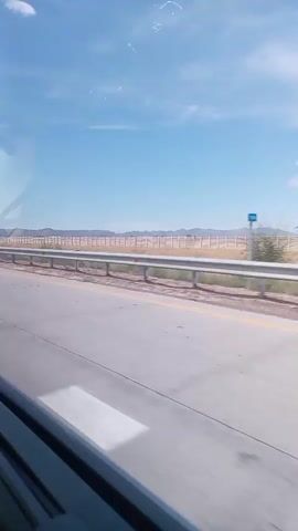 A video captures a landscape from a moving vehicle. The foreground shows a portion of a road, with white lane markings visible. A silver guardrail runs along the side of the road. Beyond the road, a dry, grassy field stretches out, dotted with yellow flowers. A long, white fence, likely a property line or enclosure, extends across the mid-ground. In the distance, mountains are visible under a clear blue sky with a few wispy clouds. The overall impression is one of a wide-open, rural or semi-rural environment.