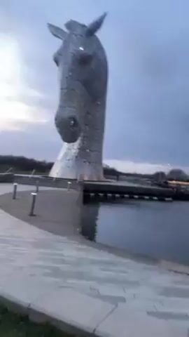 The video shows a pair of colossal horse head sculptures, named The Kelpies, standing in a body of water. They are made of shiny, silver-colored metal and are situated on platforms within the water. The horse heads are designed with an intricate, fragmented appearance. The sky is overcast, suggesting either early morning or late evening. The sculptures are surrounded by a paved walkway and a grassy area. There are a few small lights along the walkway. The video appears to be taken from a moving perspective, panning to reveal both horse heads from a close vantage point.