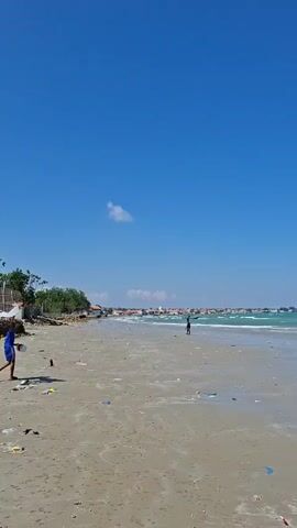 The video shows a wide sandy beach under a clear blue sky with a few wispy clouds. In the foreground, there is scattered trash on the sand, including plastic bags and other debris. To the left, a young person wearing a blue shirt and dark shorts is standing and appears to be holding a small object. Further down the beach, another person, appearing to be a child, is walking towards the water. In the middle ground, the turquoise ocean has gentle waves, and several boats are visible in the distance. Some of these boats appear to be traditional fishing vessels with canopies. Along the shoreline, there are some trees and buildings that suggest a coastal settlement. The overall impression is a somewhat neglected beach with evidence of human activity and natural beauty.