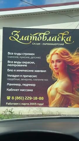 The video shows the exterior of a salon and barbershop called “Zlatovlaska” (Златовласка). First, there's a close-up of a large advertisement featuring a digital illustration of a woman with flowing golden hair, text that includes the salon's name and the services it offers (haircuts, coloring, perms, styling, manicures, pedicures, massage). The ad also lists a phone number and states that the salon has been operating since March 2003. The camera then pans up and zooms out to reveal the front of the building. It's a single-story, light-colored building with brown window frames and door. There is a small brown awning above the entrance with the salon’s name printed on it. The entrance has a few steps leading up to the door. A green metal cart with a yellow bucket is placed near the entrance. The sky is visible, with blue patches and some white clouds. The overall impression is of a small, local business.