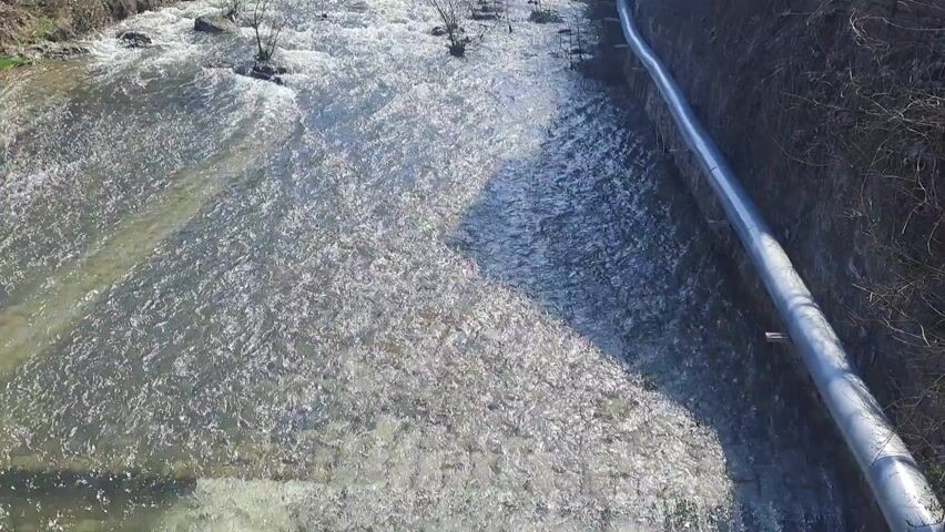 The video displays a fast-flowing river, viewed from above. The water is turbulent and appears shallow, with sunlight glinting off its surface, creating bright reflections and distinct shadows.

The river flows between concrete-lined banks on both sides. Along the right bank, a large, grey pipe runs horizontally, paralleling the river's course. In the middle of the river, several small clusters of bare, wintery bushes or young trees are visible, growing directly within the riverbed, resisting the current.

The scene is brightly lit by natural sunlight, suggesting a clear day. No people or animals are visible. The overall impression is one of a managed or channeled river in an outdoor, possibly urban or semi-urban, environment, consistent with a location like Varshets, Bulgaria. There is no visible text.