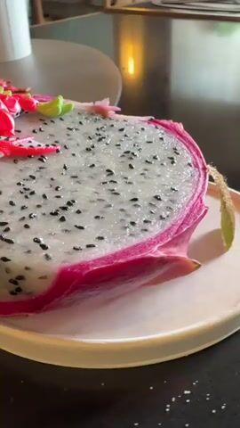 A close-up video shows a slice of dragon fruit with a pink dragon figurine placed on top. The dragon fruit has white flesh speckled with black seeds and a vibrant pink rind. The dragon figurine is red with green accents and appears to be made of plastic. It is positioned on the fruit as if it is crawling on it. The background is blurred but suggests an indoor setting with a dark countertop and some metallic surfaces. The video is filmed with a shallow depth of field, focusing on the dragon fruit and figurine.