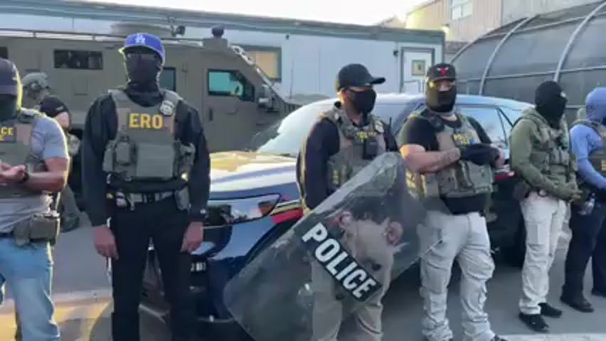 A group of approximately 10-15 individuals, mostly men, stand in formation outdoors, appearing to be law enforcement or security personnel. Most are wearing tactical gear, including bulletproof vests, balaclavas or face coverings, and some wear hats. Visible text on their vests includes "ERO" (Enforcement and Removal Operations), "POLICE ICE," and "POLICE." One individual holds a clear riot shield with "POLICE" written on it.

In the background, a dark SUV with its headlights on is visible, along with a larger armored vehicle. Further back, there's a building and a fence topped with barbed wire, suggesting a secure facility such as a detention center. The scene is illuminated by the low light of late afternoon or early evening, with the sun casting a warm glow.

An audible voice can be heard, likely from a protester, speaking to the group. Phrases such as "despicable cowards," "helpless people," and "We are not going inside the building full of helpless people" are discernible, indicating a confrontational situation, likely a protest against the officers or the facility. The individuals in tactical gear stand still and appear impassive.