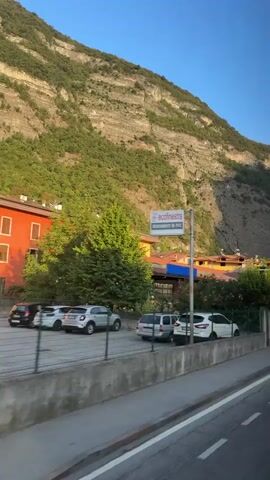 The video shows a view from inside a vehicle, looking out at a street lined with buildings and a large, vegetated mountain in the background. The sun is shining, casting long shadows. On the left side of the street, there are several cars parked and a sign that reads "ecofinestra". Further down the street, there are more buildings, some with green walls and red roofs. There is a pedestrian crossing marked on the road. The mountain behind the buildings is covered in green foliage and appears to be a steep incline with visible rock formations. The sky is clear and blue.