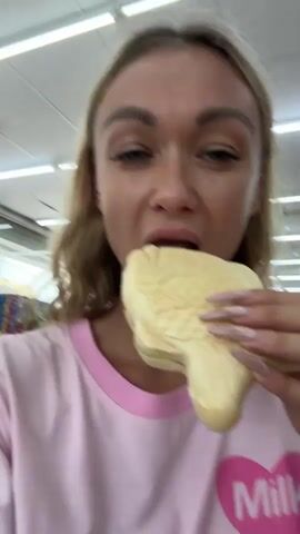 A fair-skinned woman with blond hair and manicured nails is taking a bite of a fish-shaped pastry. She is wearing a pink shirt and is indoors, presumably in a shop, as red lanterns can be seen hanging from the ceiling in the background.  The pastry is light-colored with a fish-scale pattern and appears to have a green and red filling. She seems to be recording herself as she eats.