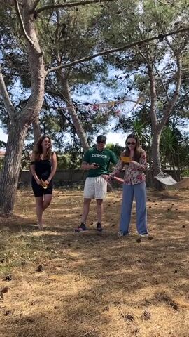 Three people are in a backyard, under pine trees. The ground is covered in dry pine needles and cones. The woman on the left wears a black top and shorts. She holds a microphone and laughs. The man in the middle wears a green Adidas shirt and white shorts and looks at his phone. The woman on the right wears a floral shirt and blue pants. She holds a yellow paper. They are celebrating International Night. There is a hammock in the background, hung between two palm trees.