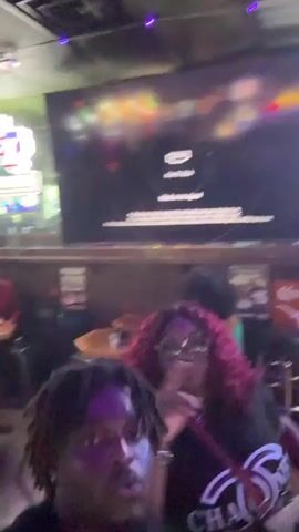 The video shows a man with dreadlocks taking a selfie video in what appears to be a bar. The man is wearing a black shirt. Behind him, a woman with red hair is seen making a peace sign, and there are other patrons and bar staff present in the background. There is a partially visible pool table. The bar is decorated with neon signs, including UFC and a Texas sign, as well as beer signs.  Several TVs are mounted on the walls, one displaying a "no signal" message. There are plates with food, and drinks on the tables. The overall atmosphere seems lively with music playing.