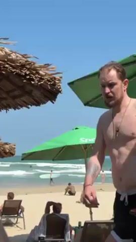 The video shows a man standing shirtless on a beach. He has light skin, a short beard, and a tattoo on his left arm. He is wearing dark shorts and a necklace.  He is surrounded by beach umbrellas, some with thatched roofs, and beach chairs.  In the background, the ocean can be seen with a person on a surfboard. The sky is blue and clear. The camera appears to be held by someone seated on a beach chair. The man bends forward, perhaps adjusting something on a beach chair.