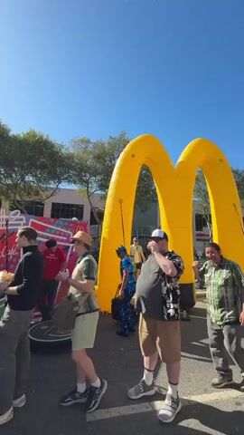 The video shows an outdoor event with a McDonald's McCafe booth and a large inflatable McDonald's arch. Several people are standing in line and milling around. The sun is shining brightly, casting shadows. The overall atmosphere appears to be festive.