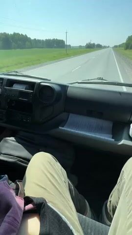 The video shows the interior of a vehicle from the perspective of a passenger, looking forward through the windshield. The dashboard is visible in the foreground, with a black console featuring buttons and vents. A white cup is placed on a ledge on the right side of the console. Papers are neatly stacked in a compartment on the dashboard. The legs of at least two people are visible in the lower portion of the frame, suggesting they are sitting in the front seats. Outside the vehicle, a paved road stretches into the distance, flanked by green fields and trees under a clear blue sky. Utility poles line the side of the road. The overall scene suggests a daytime journey on a rural highway.