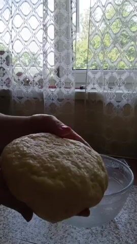 The video shows a pair of hands holding a ball of dough over a clear glass bowl filled with water. The hands then lower the dough into the water and begin to knead it. The background is blurry but appears to be a kitchen with a window covered by a lace curtain. The lighting suggests it is daytime.