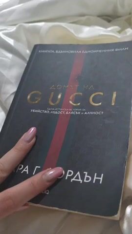 A person with manicured nails opens a book titled "The House of Gucci: A Sensational Story of Murder, Madness, Glamour, and Greed," revealing page 119. The book is laid on a shiny, white fabric, possibly satin or silk. The text on the page is in Bulgarian. The video seems to focus on presenting the book and its content.