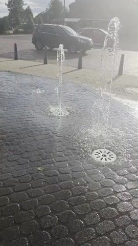 The video shows a close-up, low-angle view of a series of water jets erupting from circular grates embedded in a dark, wet cobblestone surface. The water shoots upwards in thin streams, creating splashes. In the background, out of focus, there is a paved area with bollards, a parked dark SUV, and some buildings and trees. The lighting suggests it is daytime, possibly with bright sunlight causing a hazy effect. The overall impression is of an outdoor public space with a water feature.