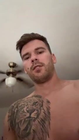 A man with short brown hair and stubble is looking into the camera, speaking. He has a large lion tattoo on his chest and shoulder. Behind him, a ceiling fan with a light fixture is visible. The man is shirtless.