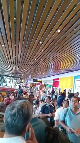 The video presents a busy scene within what appears to be an airport terminal. Many people are visible, some seated in waiting areas, others walking through the space. An airport employee, identifiable by their uniform and lanyard, walks purposefully through the crowd.

The environment features modern architectural design, particularly in the ceiling which is composed of numerous parallel wooden or metal slats. The walls showcase a vibrant color scheme, with segments painted in bright red and yellow.

The general atmosphere suggests a bustling public space, where people are either in transit or waiting for their flights. The video focuses on capturing the dynamics and activity of this indoor setting.