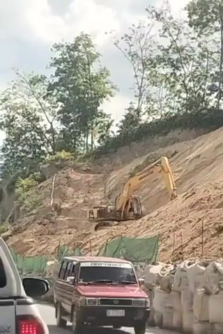 The video shows a construction site with an excavator digging into a dirt hillside. In the foreground, there is a red van driving on a road. The road is lined with sandbags and green netting, likely for erosion control. The sky is cloudy, and there are trees on the top of the hill.
