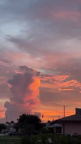 A time-lapse video shows a sunset with vibrant orange and pink clouds, gradually shifting from light to dark. Silhouetted rooftops and trees are visible at the bottom of the frame, with power lines running horizontally across the sky. The clouds are thick and textured, with some areas appearing brighter and more intense than others. The overall impression is one of a dramatic and beautiful natural phenomenon.