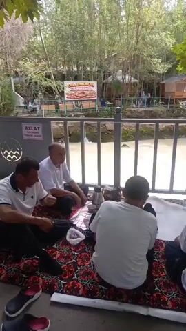 Four men are sitting on the ground, on colorful patterned blankets, next to a metal fence, with a river visible beyond the fence. Two of the men are facing the camera, while the other two are facing away from the camera. One man in the foreground, facing the camera, is wearing a white polo shirt and is holding a knife in his right hand, appearing to cut something on a red and white checkered cloth spread on the ground. Another man, also facing the camera, is wearing a white shirt and white cap, and is also sitting on the ground. Behind them, two men are seated with their backs to the camera. One of them is wearing a white t-shirt, and the other is wearing a light-colored shirt. In the background, beyond the river, there are trees and several wooden structures. A sign with writing and images is visible on a structure in the background. There are also some leaves on the ground in the foreground. There appears to be a thermos or jug on the ground between the men.