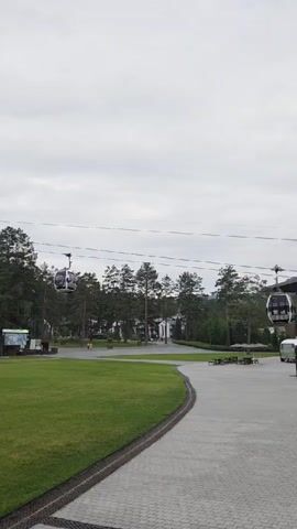 A person is walking on a paved path in front of a manicured lawn. There are three cable cars suspended on wires above the scene. One cable car is to the left of the frame, and two are to the right, moving from left to right. Trees line the background. The sky is overcast.