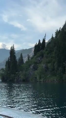 The video captures a scenic view from a boat on a lake. The water is dark and rippling, reflecting the sky and surrounding landscape. Towering mountains, some with snow-capped peaks, dominate the background. Lush green trees cover the slopes of the closer mountains, while the distant ones fade into a hazy blue. The sky is partly cloudy with sunlight breaking through, casting shadows and highlighting the mountainous terrain. The front of the boat is visible in the foreground, with a rope and some metal fixtures. The camera pans slowly to the right, revealing more of the expansive mountain range and the tranquil lake.